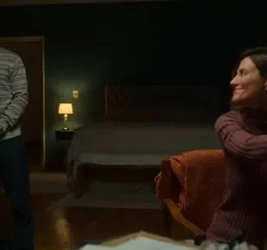 Homem sorridente na porta de um quarto com pouca luz, olhando para mulher sentada que sorri. Cena doméstica e acolhedora.