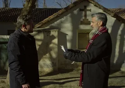 Dois homens em frente a uma capela rústica com uma cruz. Um de barba e cachecol lê papéis, o outro observa. Clima sério.