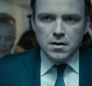 Homem de terno com olhar intenso em primeiro plano. Pessoas borradas em cenário escuro e frio. Clima de suspense.