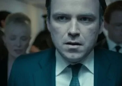 Homem de terno com olhar intenso em primeiro plano. Pessoas borradas em cenário escuro e frio. Clima de suspense.