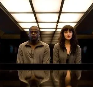 Um homem e uma mulher, sérios, sentados em sala escura e futurista. Teto iluminado em grade e telas azuis/roxas nas paredes.