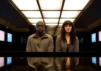 Um homem e uma mulher, sérios, sentados em sala escura e futurista. Teto iluminado em grade e telas azuis/roxas nas paredes.