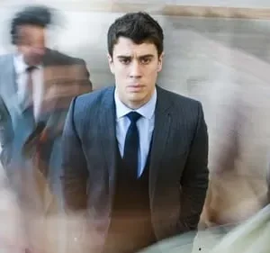 Jovem de terno escuro em foco, expressão séria. Figuras borradas passam rapidamente, criando um clima de urgência e isolamento.