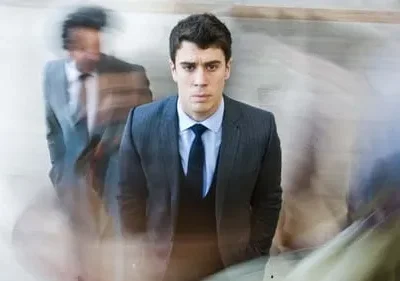 Jovem de terno escuro em foco, expressão séria. Figuras borradas passam rapidamente, criando um clima de urgência e isolamento.