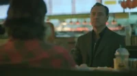 Homem sério de terno escuro senta-se à mesa de uma lanchonete, olhando fixamente. Pessoa de camisa xadrez de costas no primeiro plano.