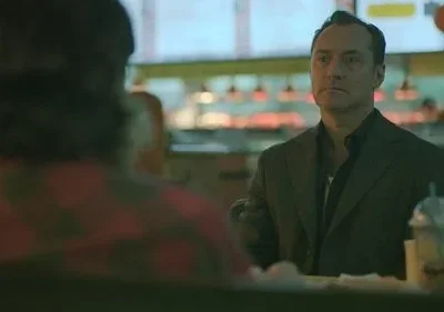 Homem sério de terno escuro senta-se à mesa de uma lanchonete, olhando fixamente. Pessoa de camisa xadrez de costas no primeiro plano.