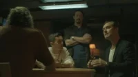 Quatro homens em uma sala escura. Dois à mesa, um gesticula. Um homem de braços cruzados no fundo. Atmosfera séria.