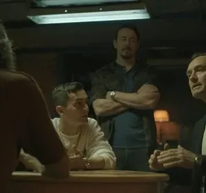 Quatro homens em uma sala escura. Dois à mesa, um gesticula. Um homem de braços cruzados no fundo. Atmosfera séria.