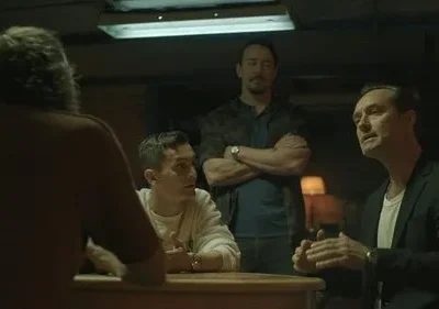 Quatro homens em uma sala escura. Dois à mesa, um gesticula. Um homem de braços cruzados no fundo. Atmosfera séria.