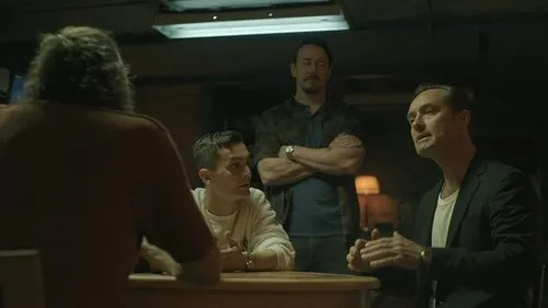 Quatro homens em uma sala escura. Dois à mesa,um gesticula. Um homem de braços cruzados no fundo. Atmosfera séria.