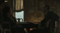 Um homem e uma mulher se encaram em uma mesa em um quarto escuro. A luz ambiente destaca seus perfis, criando um clima tenso e introspectivo.