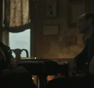 Um homem e uma mulher se encaram em uma mesa em um quarto escuro. A luz ambiente destaca seus perfis, criando um clima tenso e introspectivo.