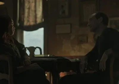 Um homem e uma mulher se encaram em uma mesa em um quarto escuro. A luz ambiente destaca seus perfis, criando um clima tenso e introspectivo.