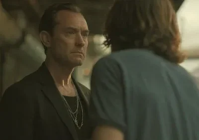 Homem sério com casaco escuro e colares olha para pessoa de costas com cabelo longo. Cenário urbano com iluminação discreta.