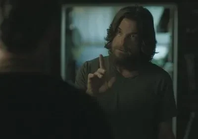 Um homem barbudo de cabelo comprido aponta o dedo para outro homem de costas, em um ambiente escuro e sério.