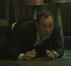 Homem de terno escuro rasteja no chão, olhando assustado para a frente em um ambiente escuro com colunas metálicas.