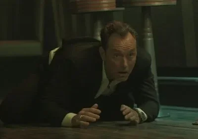 Homem de terno escuro rasteja no chão, olhando assustado para a frente em um ambiente escuro com colunas metálicas.