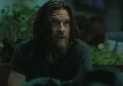 Homem barbudo de cabelo comprido e olhos arregalados, com expressão intensa, em quarto escuro com plantas verdes.