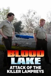 Dois homens de uniforme cáqui em pé na margção de um lago,com um barco salva-vidas. Fundo com árvores secas,clima sério.