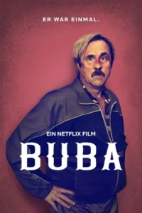 Homem com bigode,nariz machucado e cabelo loiro,usando agasalho,posa confiantemente em fundo vermelho.