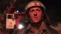 Jovem bombeiro com capacete e estrela vermelha, rosto avermelhado de sujeira, expressão séria. Caminhão de bombeiros ao fundo. Cena noturna e tensa.