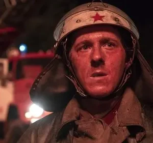 Jovem bombeiro com capacete e estrela vermelha, rosto avermelhado de sujeira, expressão séria. Caminhão de bombeiros ao fundo. Cena noturna e tensa.