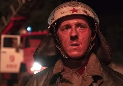 Jovem bombeiro com capacete e estrela vermelha, rosto avermelhado de sujeira, expressão séria. Caminhão de bombeiros ao fundo. Cena noturna e tensa.