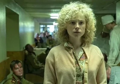 Mulher de cabelo loiro encaracolado, expressão preocupada, em corredor de hospital com vários pacientes e pessoal médico. Cenário tenso.