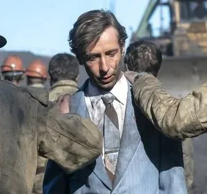 Um homem pálido de terno sujo é amparado por trabalhadores empoeirados num canteiro industrial, com trator ao fundo. Clima sombrio.