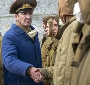 Oficial de uniforme azul e quepe aperta a mão de homem em traje protetor. Fila de figuras em cáqui ao fundo. Clima sombrio.
