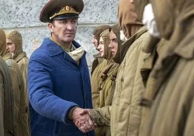 Oficial de uniforme azul e quepe aperta a mão de homem em traje protetor. Fila de figuras em cáqui ao fundo. Clima sombrio.