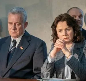 Em uma sala formal, um homem de terno com pin soviético e uma mulher de blazer xadrez observam gravemente. Outras figuras ao fundo.