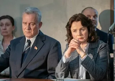 Em uma sala formal, um homem de terno com pin soviético e uma mulher de blazer xadrez observam gravemente. Outras figuras ao fundo.