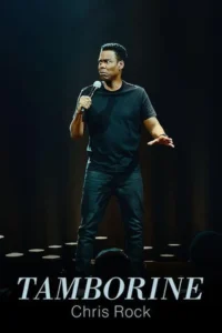 Um homem negro em pé no palco sob holofote,segurando um microfone. Ele usa roupa escura e tem uma expressão facial intensa.
