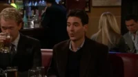 Dois homens em um bar com pouca luz. Um loiro bebe cerveja enquanto o moreno fala. Atmosfera social e descontraída.