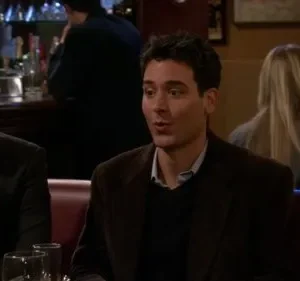 Dois homens em um bar com pouca luz. Um loiro bebe cerveja enquanto o moreno fala. Atmosfera social e descontraída.