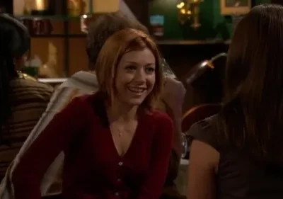 Mulher ruiva de sorriso largo conversa com amiga em um bar com luz baixa. Ambiente acolhedor e descontraído.