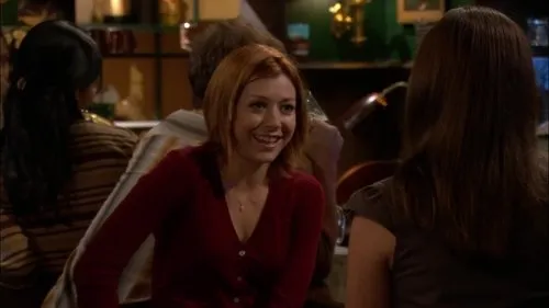 Mulher ruiva de sorriso largo conversa com amiga em um bar com luz baixa. Ambiente acolhedor e descontraído.