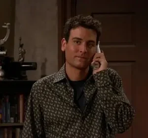 Jovem com camisa estampada sorri, falando ao celular. Ao fundo, uma estante de livros com um projetor antigo e um quadro.