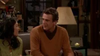 Um homem de suéter laranja conversa com uma mulher em um ambiente acolhedor, iluminado por uma vela e uma luminária. Clima amigável.