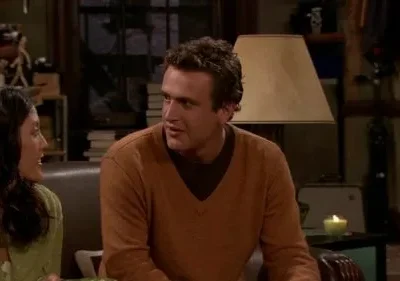 Um homem de suéter laranja conversa com uma mulher em um ambiente acolhedor, iluminado por uma vela e uma luminária. Clima amigável.