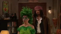Duas pessoas fantasiadas: uma de pássaro verde emplumado com maquiagem elaborada, outra de pirata barbudo. Ambiente festivo e aconchegante.