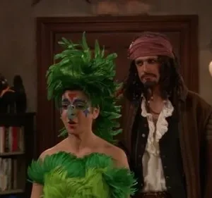 Duas pessoas fantasiadas: uma de pássaro verde emplumado com maquiagem elaborada, outra de pirata barbudo. Ambiente festivo e aconchegante.