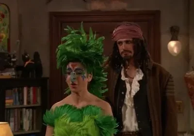 Duas pessoas fantasiadas: uma de pássaro verde emplumado com maquiagem elaborada, outra de pirata barbudo. Ambiente festivo e aconchegante.