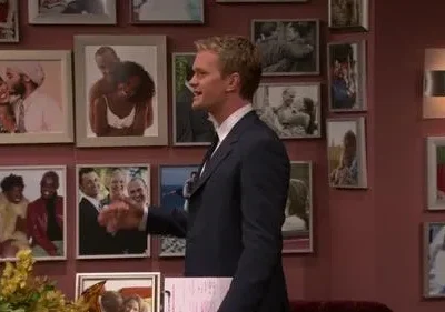 Homem loiro de terno sorri, gesticula. Parede ao fundo coberta por dezenas de fotos de casais e famílias. Ambiente pessoal e divertido.