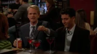 Em um bar, um homem loiro de terno gesticula para um moreno sorridente. Coquetéis vermelhos sobre a mesa, ambiente noturno social.