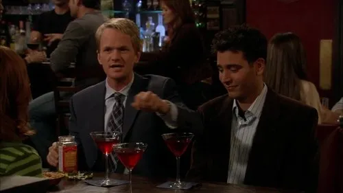 Em um bar,um homem loiro de terno gesticula para um moreno sorridente. Coquetéis vermelhos sobre a mesa,ambiente noturno social.