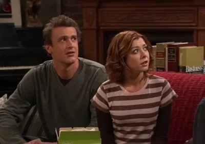 Um homem de cabelo cacheado e suéter, e uma mulher ruiva de blusa listrada, ambos surpresos e olhando para cima, em uma sala de estar.