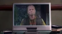 Um homem loiro, barbudo e de roupa hippie tem uma expressão emocionada em um cenário escuro e arborizado. Clima sombrio.