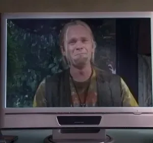 Um homem loiro, barbudo e de roupa hippie tem uma expressão emocionada em um cenário escuro e arborizado. Clima sombrio.